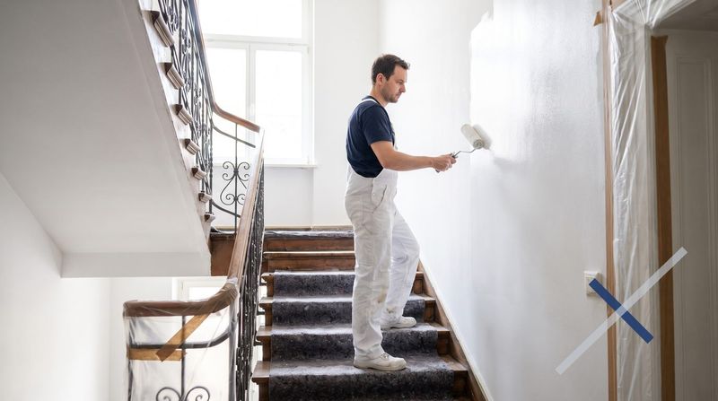 Maler streicht mit der Farbrolle eine Wand im Treppenhaus, Stufen und Geländer sind mit Folie und Malervlies abgedeckt